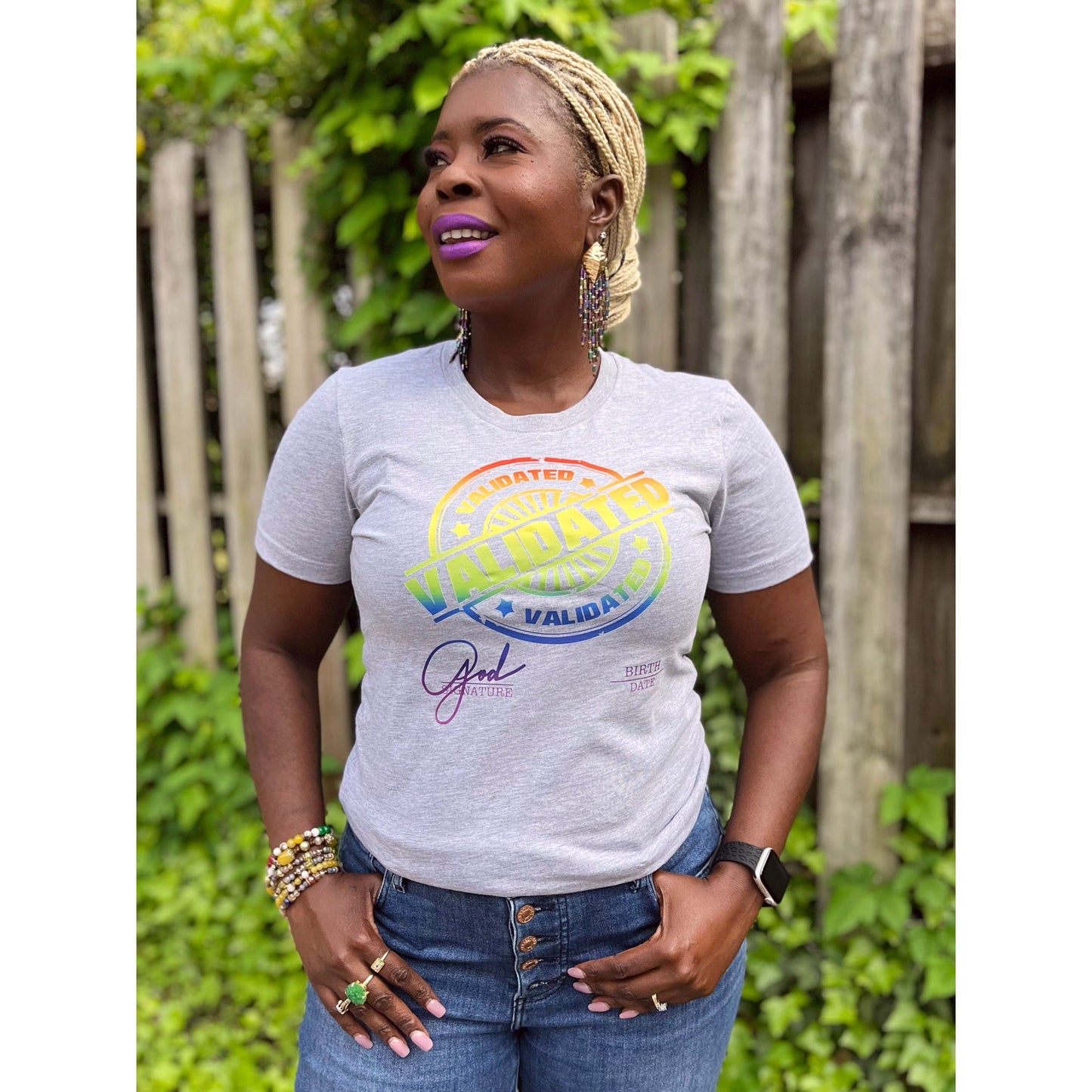 Smiling Black woman standing outdoors in a heather gray “Validated” tee with a rainbow stamp design, layered bracelets, and Mardi Gras Peacock dangle earrings.