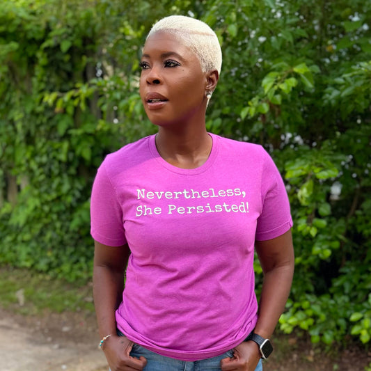 Person wearing a pink t-shirt with 'Nevertheless, She Persisted!' text outdoors.