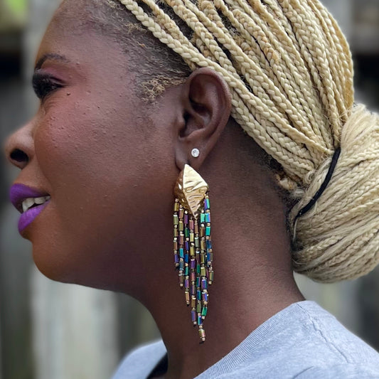 Smiling Black woman standing outdoors in a heather gray “Validated” tee with a rainbow stamp design, layered bracelets, and Mardi Gras Peacock dangle earrings.