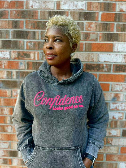 Person wearing a hoodie with 'Confidence looks good on me' text against a brick wall.