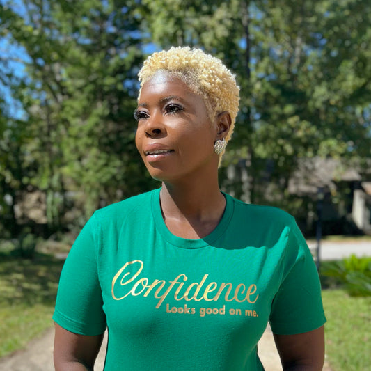 Woman wearing a kelly green t-shirt with 'Confidence looks good on me' text in metallic gold, outdoors.
