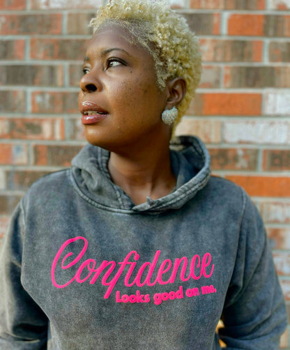 Person wearing a hoodie with 'Confidence Looks good on me' text against a brick wall.