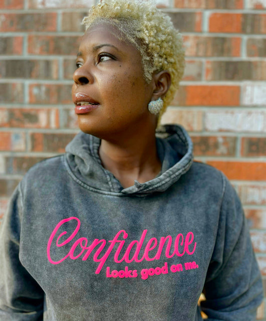 Person wearing a hoodie with 'Confidence Looks good on me' text against a brick wall.