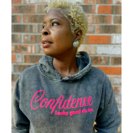 Person wearing a hoodie with 'Confidence Looks good on me' text against a brick wall.