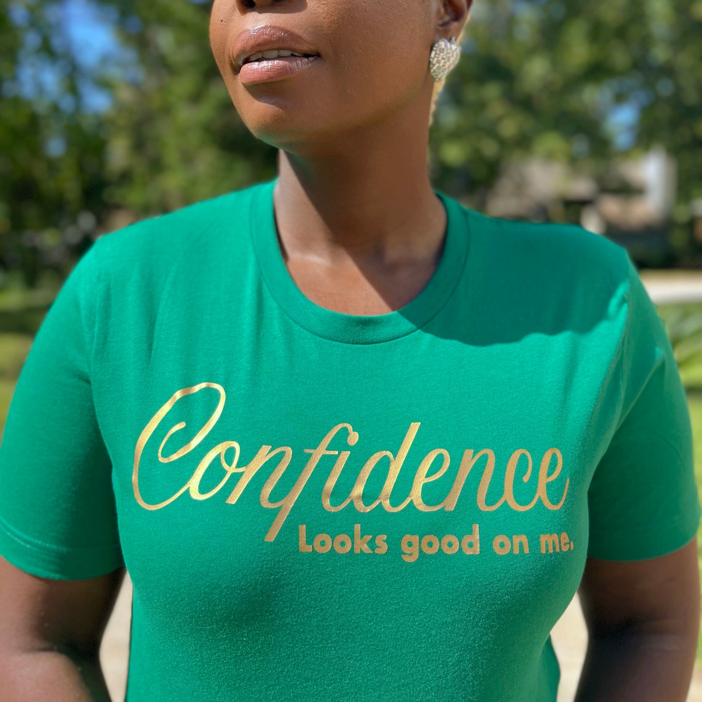 Woman wearing a kelly green t-shirt with 'Confidence looks good on me' text  in metallic gold, outdoors.