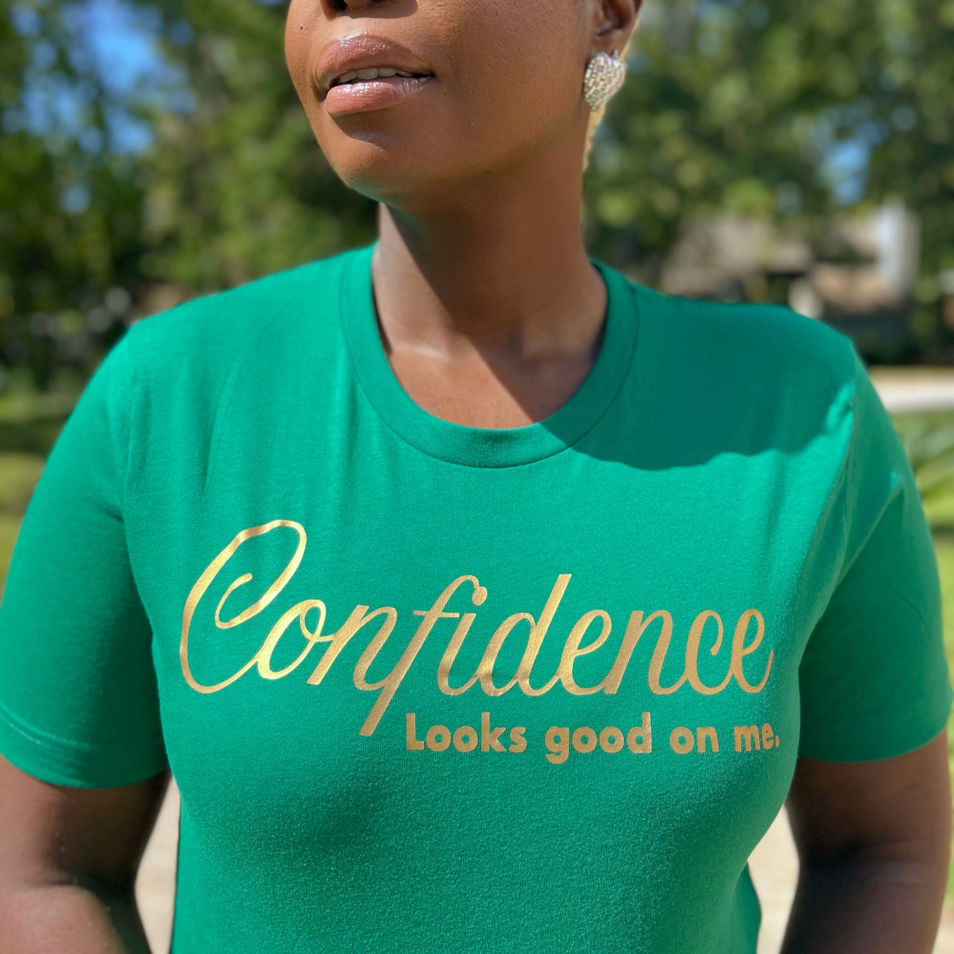 Woman wearing a kelly green t-shirt with 'Confidence looks good on me' text  in metallic gold, outdoors.