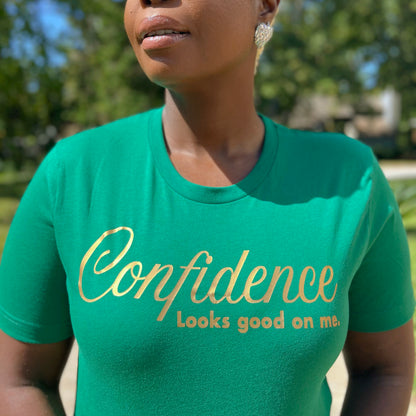 Woman wearing a kelly green t-shirt with 'Confidence looks good on me' text  in metallic gold, outdoors.