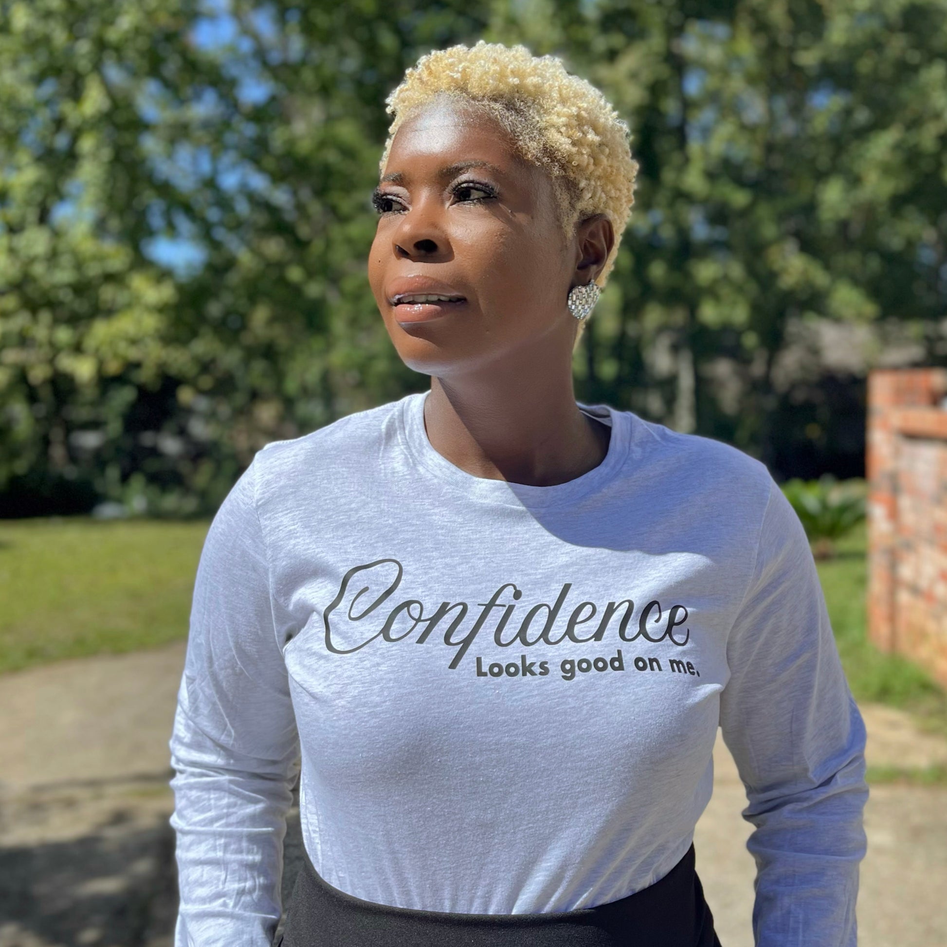 Woman wearing a ash grey long-sleeve shirt with 'Confidence Looks good on me' text in black, outdoors. 