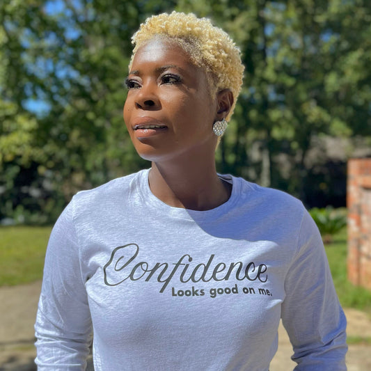 Woman wearing a ash grey long-sleeve shirt with 'Confidence Looks good on me' text in black, outdoors. 