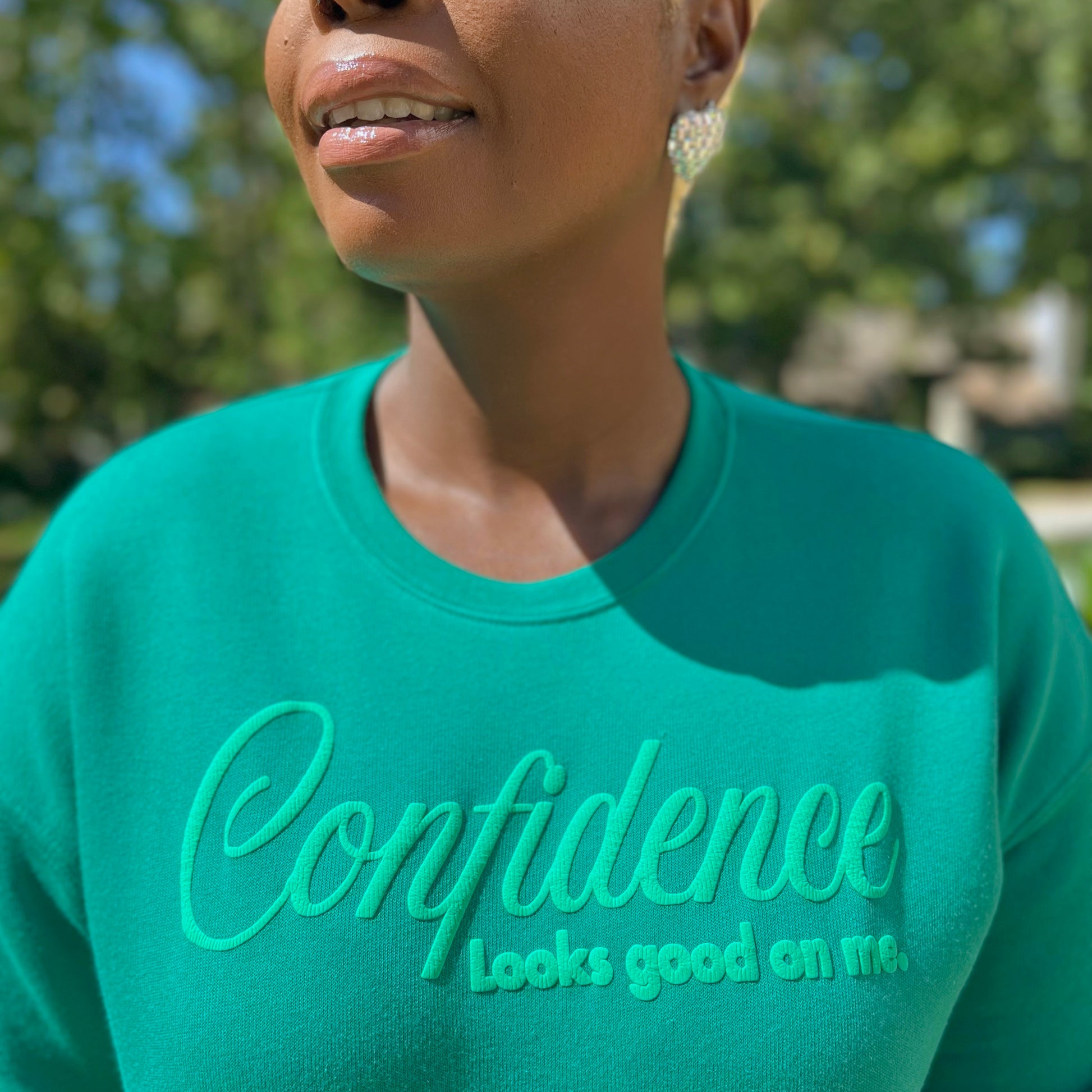 woman wearing a kelly green sweatshirt with 'Confidence looks good on me' text outdoors.
