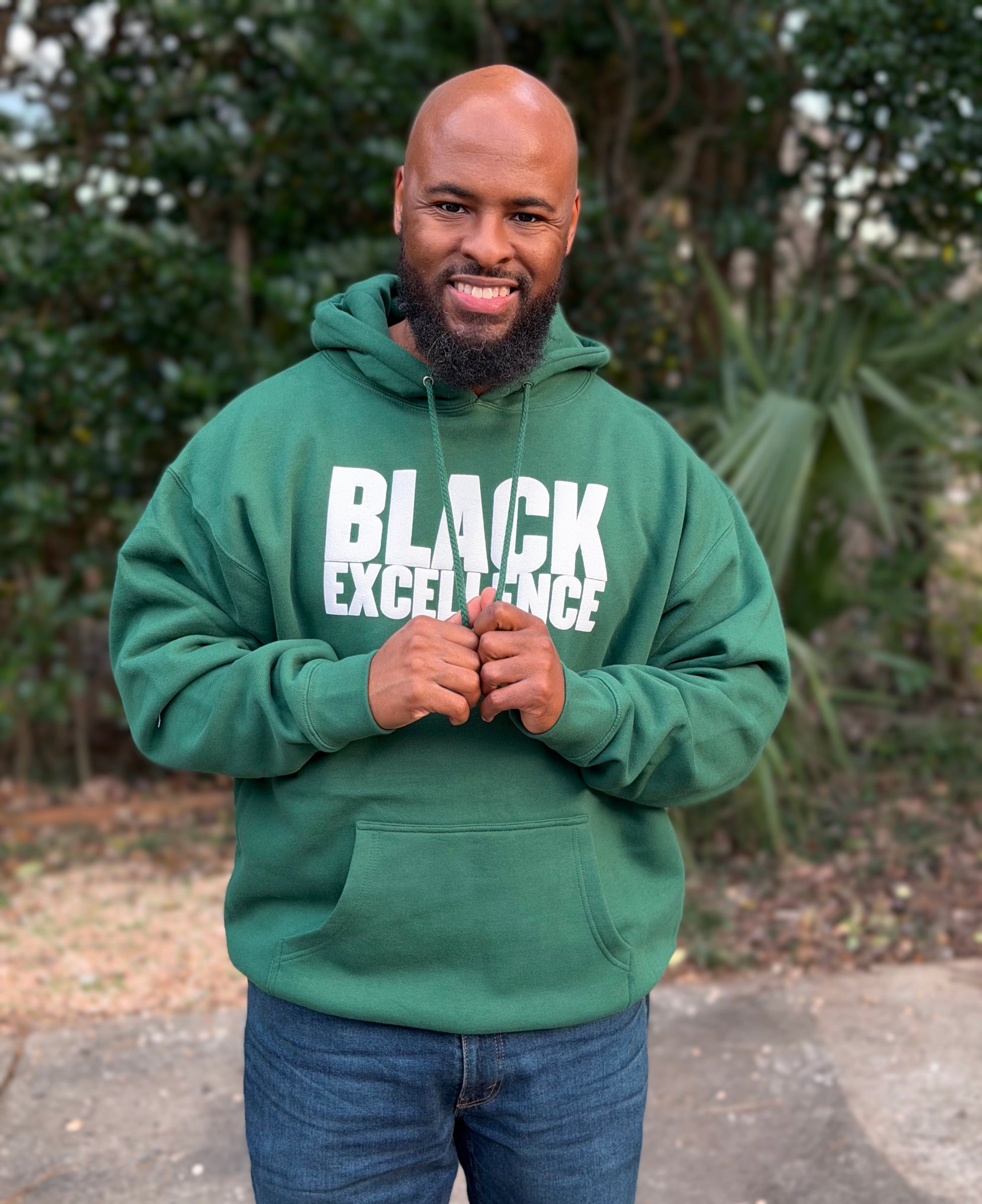 Male wearing a forest green hoodie with “Black Excellence” in bold white puff print.
