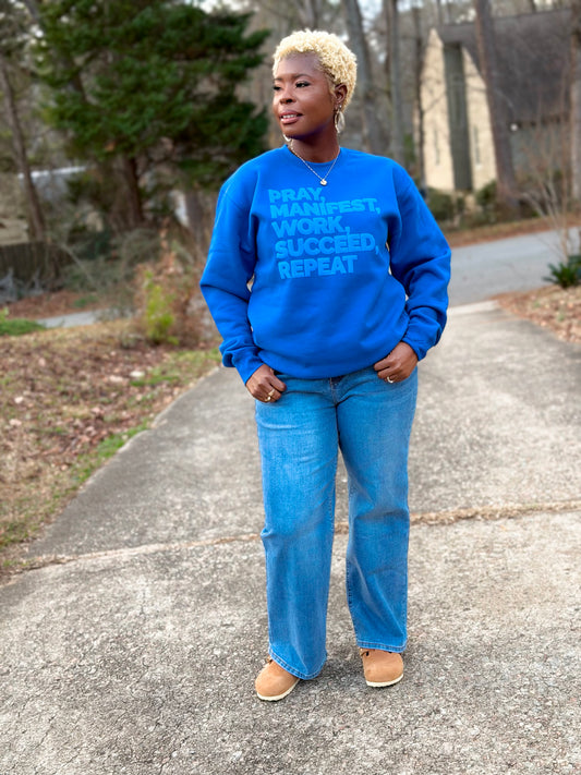 Model wearing a royal blue sweatshirt featuring the “Pray, Manifest, Work, Succeed, Repeat” design in puff print; with light blue denim and tan birkenstocks