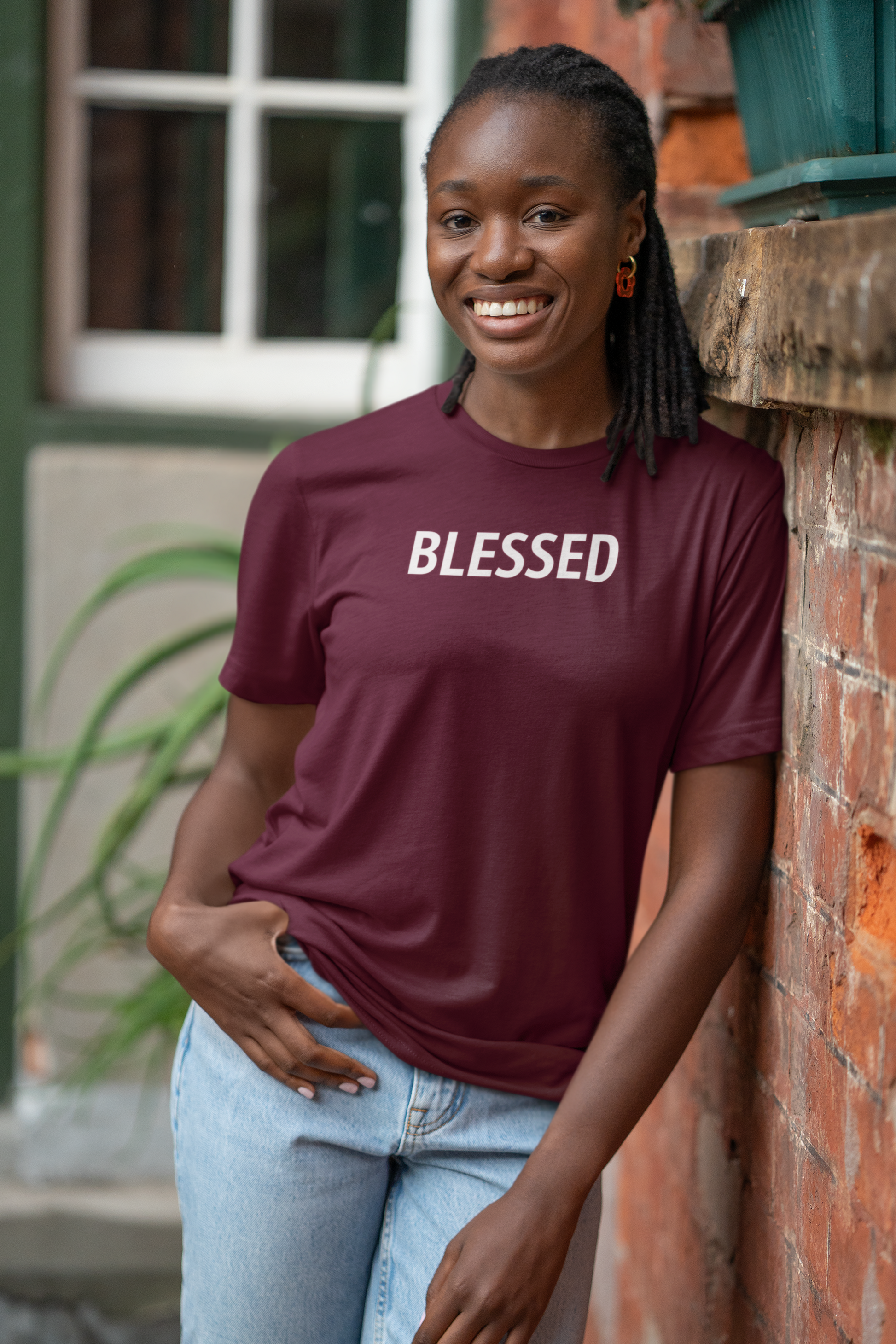A smiling woman with braided hair leans against a brick wall wearing a burgundy short-sleeve T-shirt with the word 'BLESSED' printed in white across the chest, paired with light blue jeans.