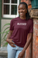 A smiling woman with braided hair leans against a brick wall wearing a burgundy short-sleeve T-shirt with the word 'BLESSED' printed in white across the chest, paired with light blue jeans.