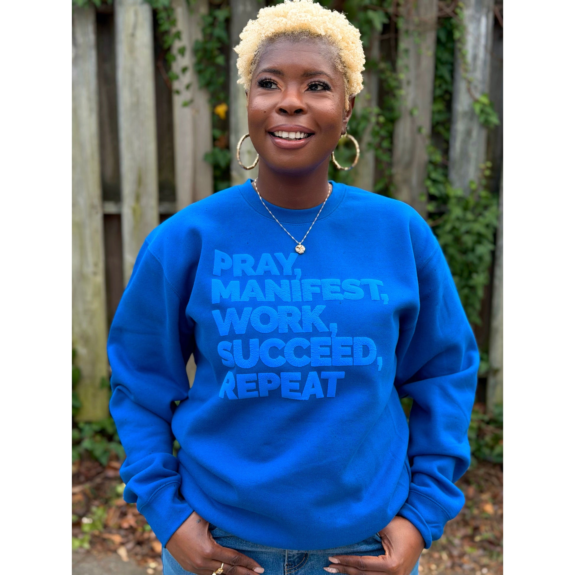 Model wearing a royal blue sweatshirt featuring the “Pray, Manifest, Work, Succeed, Repeat” design in puff print.