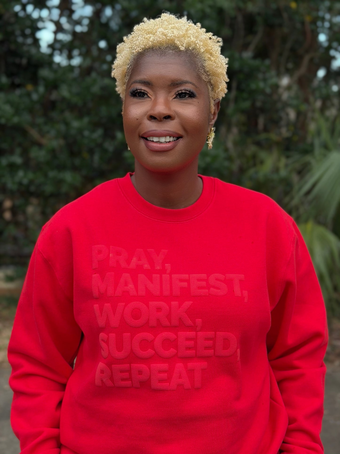 Model wearing a red sweatshirt featuring the “Pray, Manifest, Work, Succeed, Repeat” design in puff print.