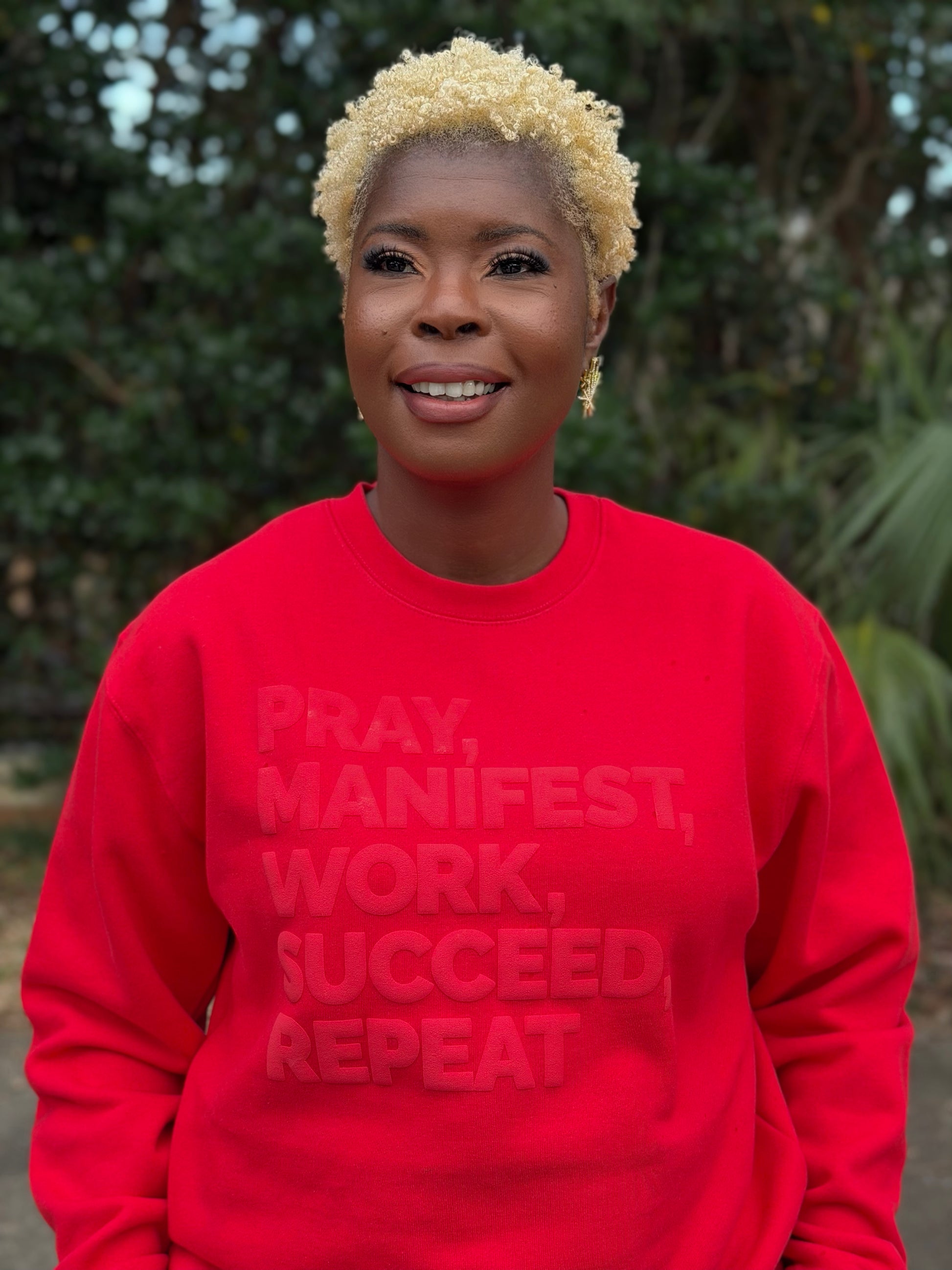 Model wearing a red sweatshirt featuring the “Pray, Manifest, Work, Succeed, Repeat” design in puff print.