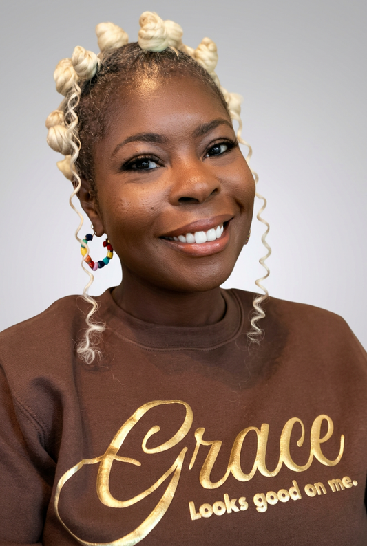 A smiling woman with blonde cornrow braids and curled ends wearing a brown sweatshirt with gold script text that reads 'Grace Looks good on me.' against a plain light gray background.