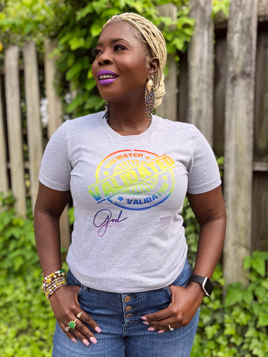 Smiling Black woman standing outdoors in a heather gray “Validated” tee with a rainbow stamp design, layered bracelets, and Mardi Gras Peacock dangle earrings.