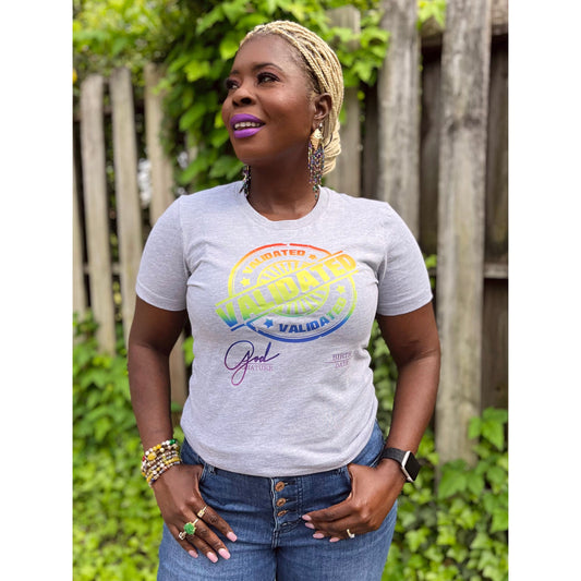 Smiling Black woman standing outdoors in a heather gray “Validated” tee with a rainbow stamp design, layered bracelets, and Mardi Gras Peacock dangle earrings.