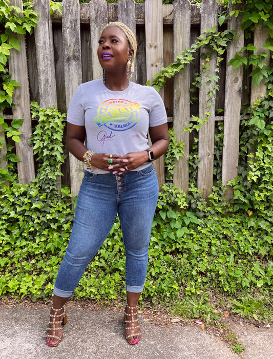 Smiling Black woman standing outdoors in a heather gray “Validated” tee with a rainbow stamp design, layered bracelets, and Mardi Gras Peacock dangle earrings.