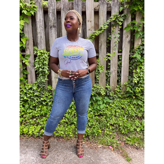 Smiling Black woman standing outdoors in a heather gray “Validated” tee with a rainbow stamp design, layered bracelets, and Mardi Gras Peacock dangle earrings.