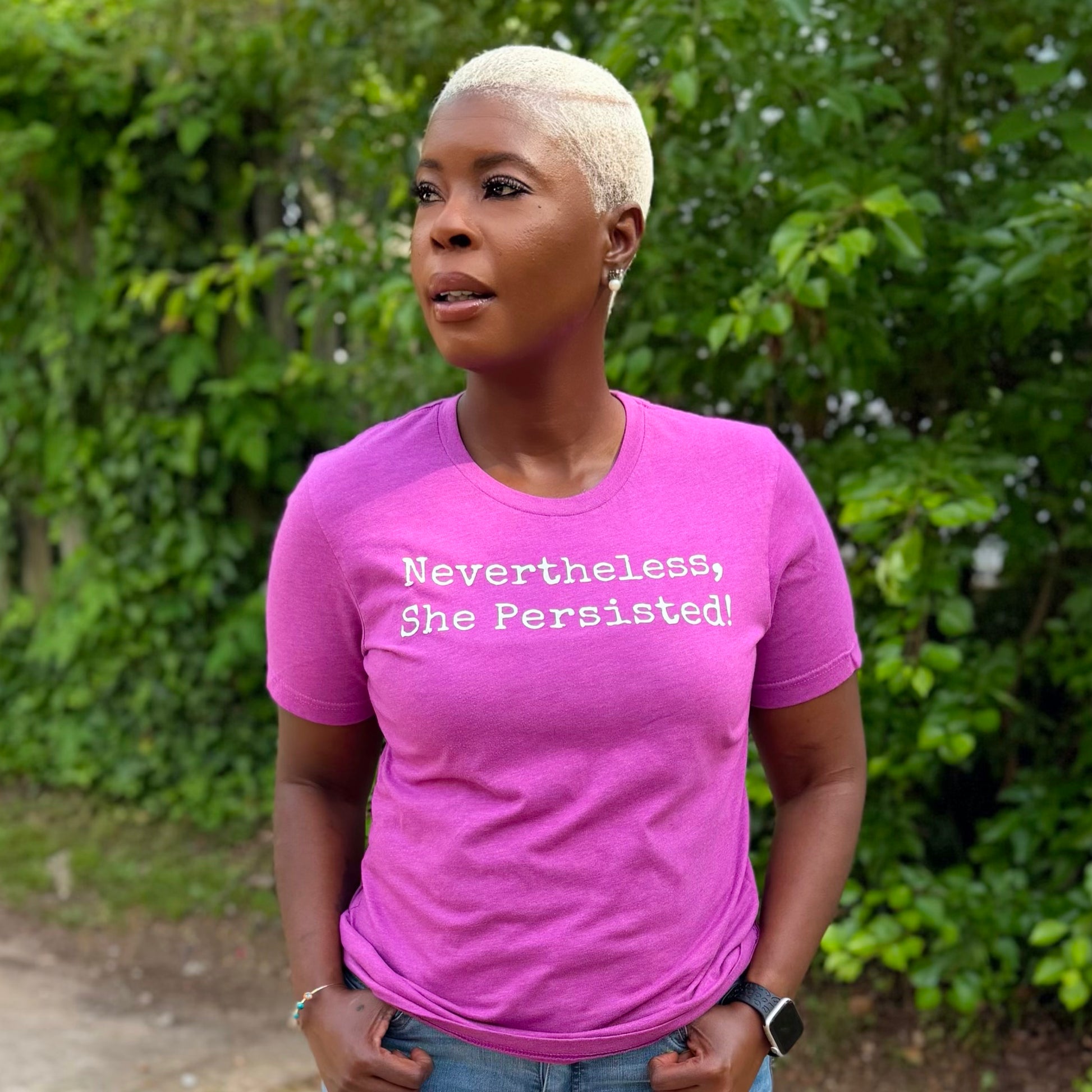 Person wearing a pink t-shirt with 'Nevertheless, She Persisted!' text outdoors.