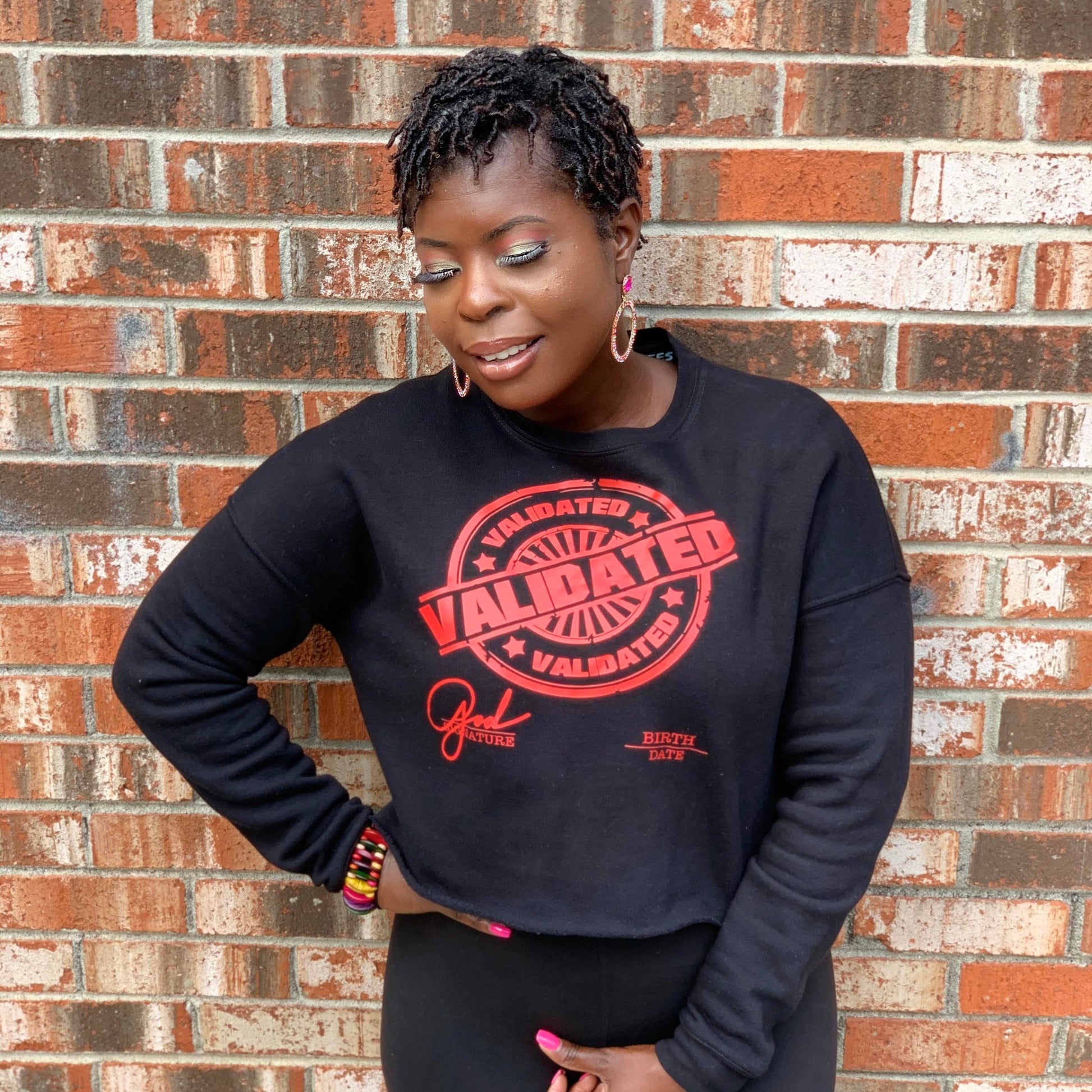 Black woman smiling confidently in a cropped black sweatshirt with a bold red “Validated” stamp graphic across the chest. Standing in front of a brick wall, one hand on her hip and the other resting near her waist.

