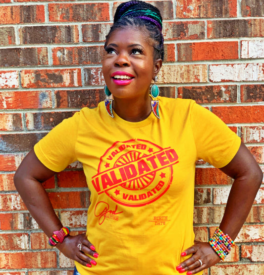 Black woman standing confidently against a brick wall in a mustard yellow “Validated” tee with red stamp design, colorful accessories, and bright pink lipstick.