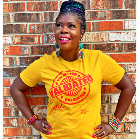 Black woman standing confidently against a brick wall in a mustard yellow “Validated” tee with red stamp design, colorful accessories, and bright pink lipstick.
